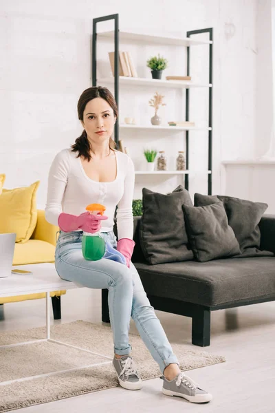 Attractive housewife looking at camera while sitting on table and holding rag and spray bottle — Stock Photo