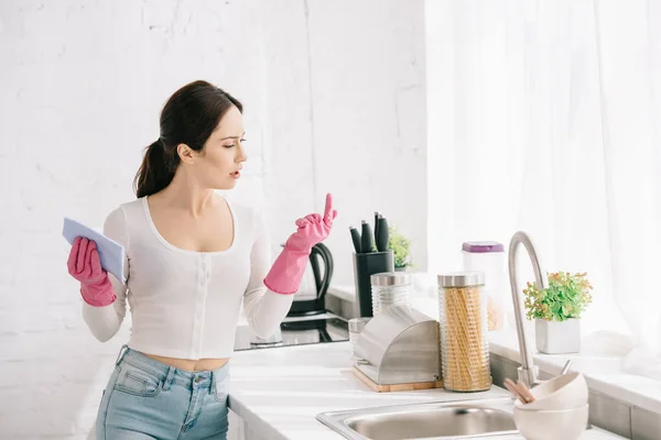 Молодая домохозяйка, стоящая на кухне в резиновых перчатках и держащая тряпку — стоковое фото