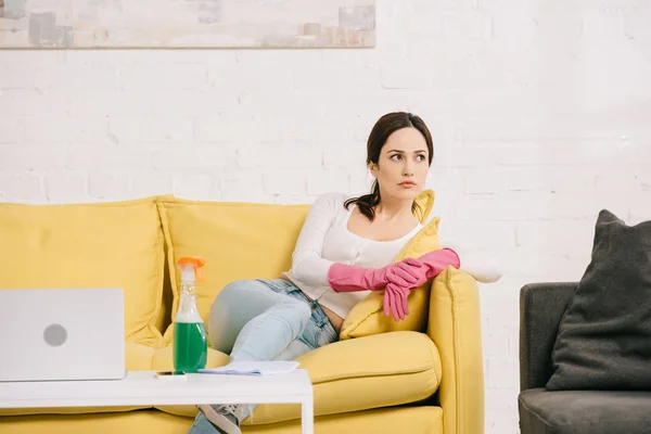 Tired housewife looking away while sitting on yellow sofa near table with laptop and spray bottle — Stock Photo