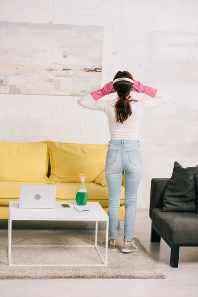 Back view of housewife in headphones standing near table with laptop and spray bottle — Stock Photo