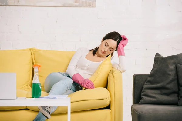 Виснажена домогосподарка сидить на жовтому дивані з закритими очима біля столу з пляшкою розпилення — стокове фото