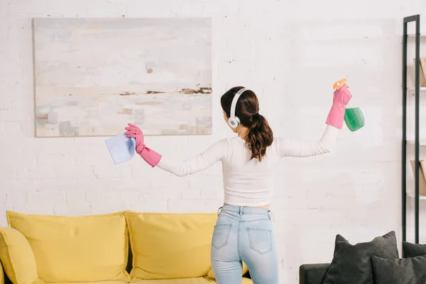 Back view of young housewife in headphones dancing while holding rag and spray bottle — Stock Photo