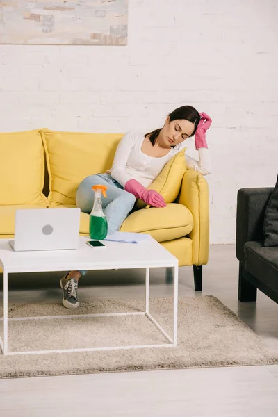 Tired housewife sitting on yellow sofa with closed eyes near table with laptop and spray bottle — Stock Photo