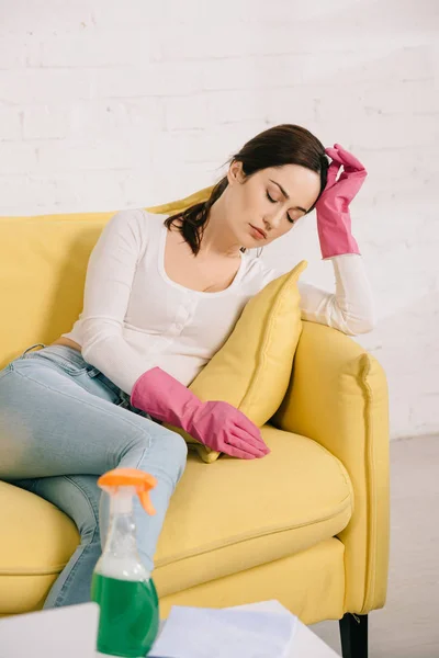 Tired housewife sitting on yellow sofa with closed eyes near table with spray bottle — Stock Photo