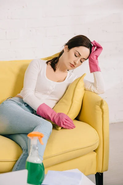 Müde Hausfrau sitzt mit geschlossenen Augen auf gelbem Sofa neben Tisch mit Sprühflasche — Stockfoto