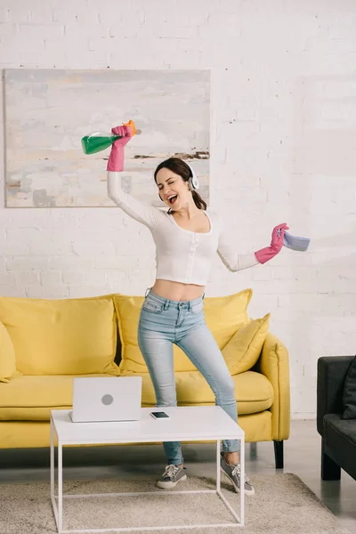 Glückliche Hausfrau mit Kopfhörern tanzt, während sie Lappen und Sprühflasche in der Hand hält — Stockfoto