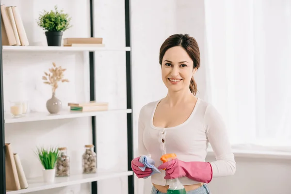 Attrayant ménagère souriant à la caméra tout en tenant chiffon et vaporisateur bouteille avec détergent — Photo de stock