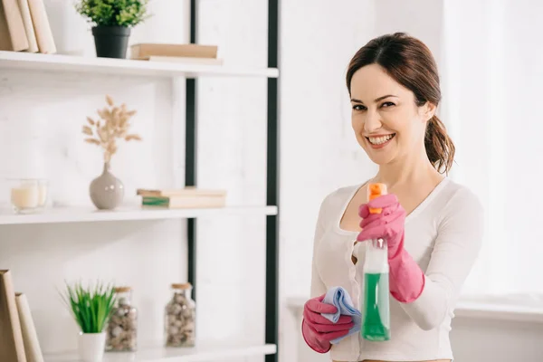 Fröhliche Hausfrau lächelt in die Kamera, während sie Lappen und Sprühflasche mit Waschmittel in der Hand hält — Stockfoto