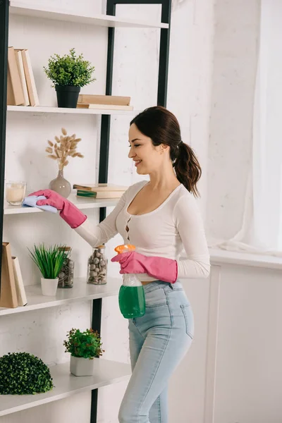 Prateleira de limpeza feliz dona de casa com pano e segurando frasco de spray com detergente — Fotografia de Stock