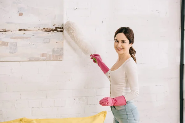Весёлая домохозяйка улыбается в камеру, когда чистит фотографию на стене пыльной щеткой — стоковое фото
