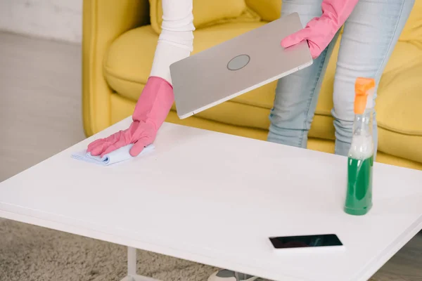 Vista ritagliata di casalinga che tiene il computer portatile mentre pulisce il tavolo con straccio — Foto stock