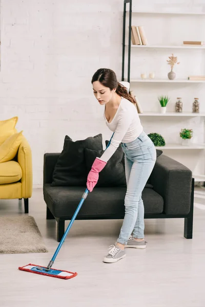 Junge Hausfrau wäscht Fußboden mit Wischmopp neben grauem Sofa — Stockfoto