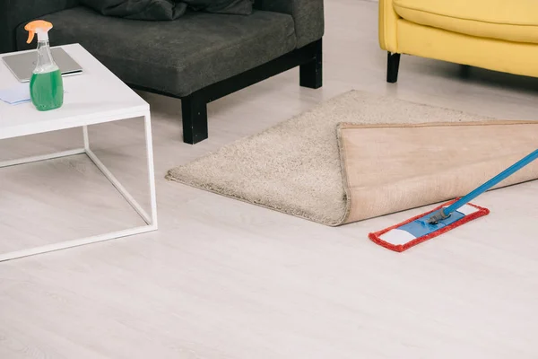 Sala de estar con sofás amarillos y grises, escritorio con botella de spray y fregona cerca de la alfombra en el suelo - foto de stock