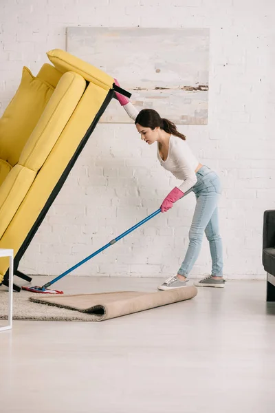 Junge Hausfrau hebt gelbes Sofa beim Fußbodenwaschen mit Wischmopp hoch — Stockfoto