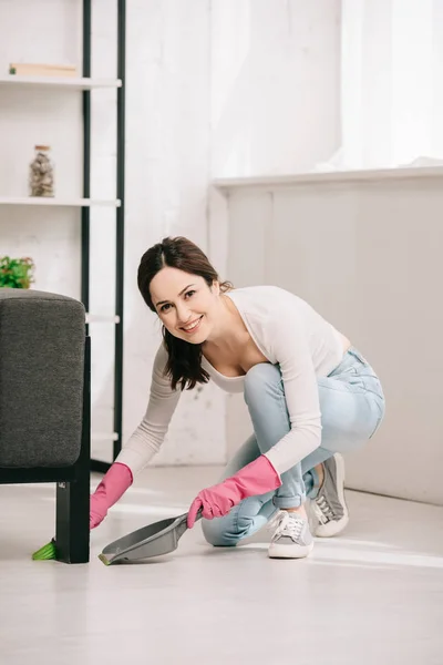 Alegre ama de casa mirando a la cámara mientras barre el suelo con cepillo y cuchara - foto de stock