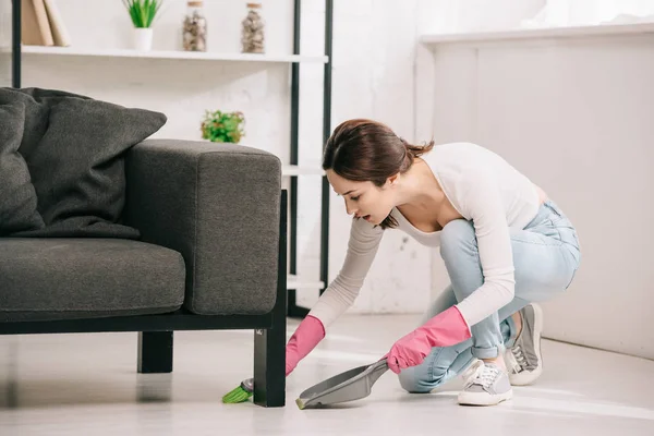 Junge Hausfrau fegt Fußboden neben Sofa mit Pinsel und Schaufel — Stockfoto