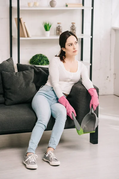 Tired housewife sitting on grey sofa and holding brush and scoop — Stock Photo