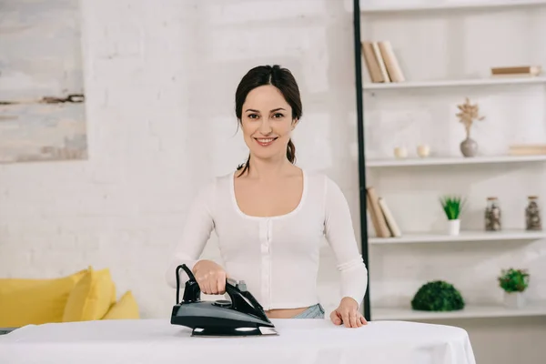 Fröhliche Hausfrau lächelt in die Kamera, während sie am Bügelbrett bügelt — Stockfoto