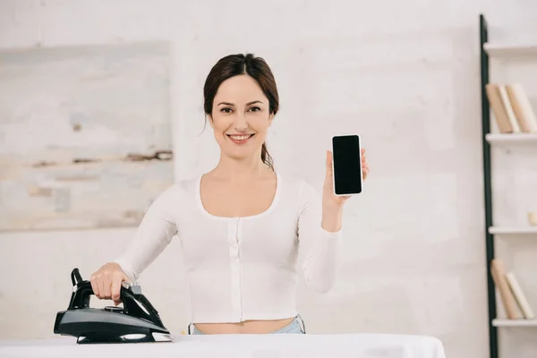 Joyeuse femme au foyer souriant à la caméra tout en se tenant près de la planche à repasser et montrant smartphone avec écran blanc — Photo de stock