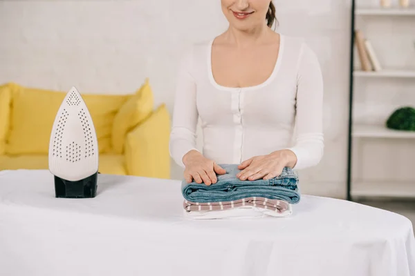 Cropped view of young housewife smiling while standing near ironing board with ironed clothes — Stock Photo