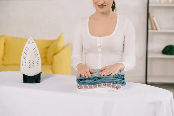 Vista recortada de la joven ama de casa sonriendo mientras está de pie cerca de tabla de planchar con ropa planchada - foto de stock