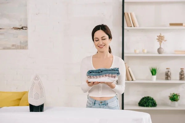 Happy housewife holding ironed clothes while standing near ironing board — Stock Photo