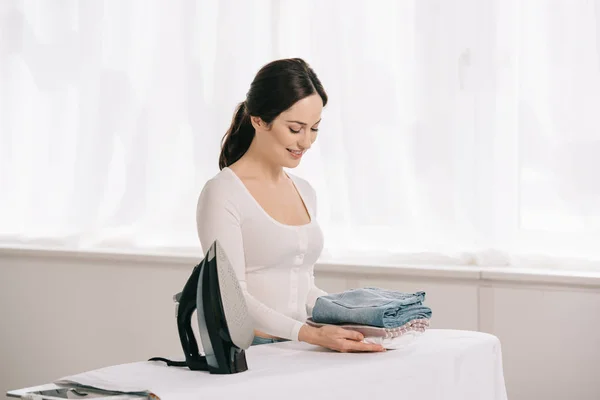 Sonriente ama de casa sosteniendo ropa planchada mientras está de pie cerca de tabla de planchar - foto de stock