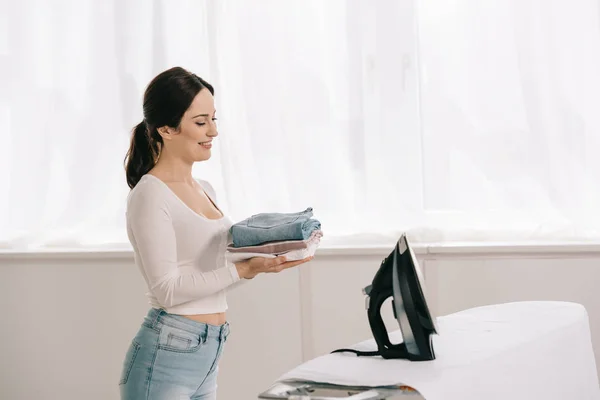 Happy housewife holding ironed clothes while standing near ironing board — Stock Photo