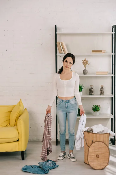 Jeune femme au foyer en jeans denim tenant des vêtements et regardant la caméra tout en se tenant près du panier à linge — Photo de stock