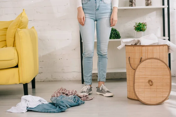 Cropped view of housewife in denim jeans standing near laundry basket and clothes on floor — Stock Photo