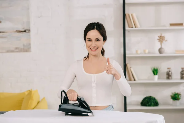 Alegre ama de casa planchado y mostrando el pulgar hacia arriba mientras mira a la cámara - foto de stock