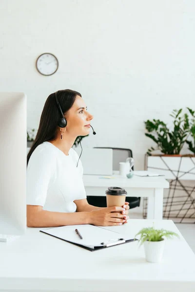 Attractive broker in headset sitting and holding paper cup in office — Stock Photo