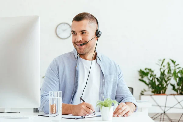 Fröhlicher Makler mit Headset, der auf Computermonitor blickt und Stift in der Hand hält — Stockfoto