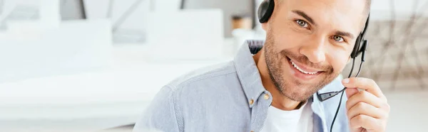 Tiro panorámico de operador feliz en la agencia de corredores tocando auriculares y mirando a la cámara - foto de stock