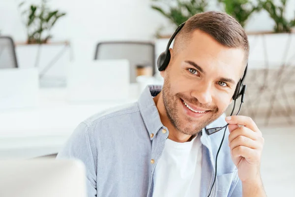 Selective focus of happy operator in brokers agency touching headset and looking at camera in office — Stock Photo