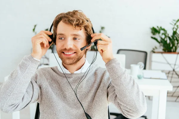 Happy bearded broker touching headset and smiling in office — Stock Photo