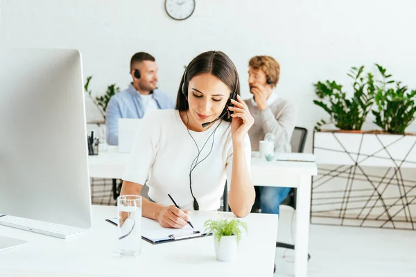 Selective focus of attractive broker touching headset and writing near coworkers — Stock Photo