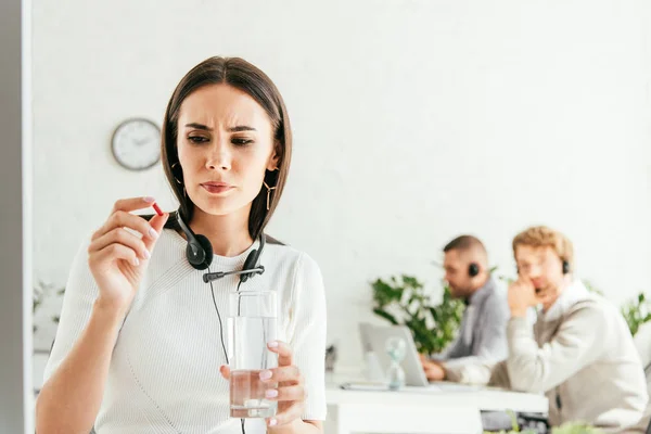 Messa a fuoco selettiva del broker malato che tiene pillola e bicchiere d'acqua vicino ai colleghi in ufficio — Foto stock