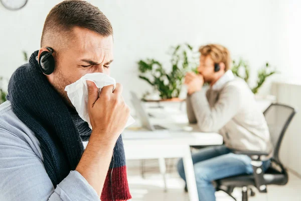 Messa a fuoco selettiva di broker malato con gli occhi chiusi starnuti nei tessuti vicino al collega in ufficio — Foto stock