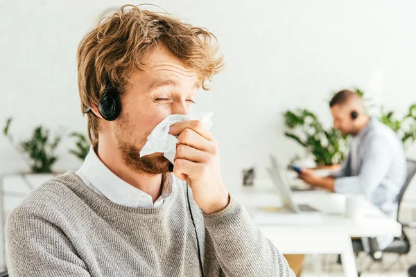 Sick bearded broker with closed eyes sneezing in tissue near coworker in office — Stock Photo