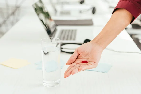 Vista cortada de corretor segurando pílula perto de vidro de água e laptop no escritório — Fotografia de Stock