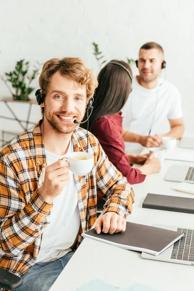 Enfoque selectivo de corredor barbudo feliz sosteniendo taza de café cerca de compañeros de trabajo - foto de stock