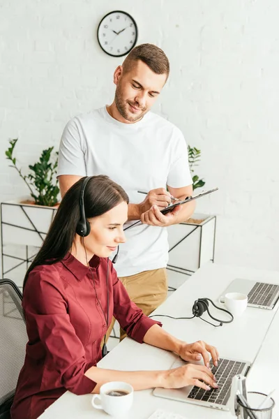 Handsome broker holding clipboard while standing near woman in headset — Stock Photo