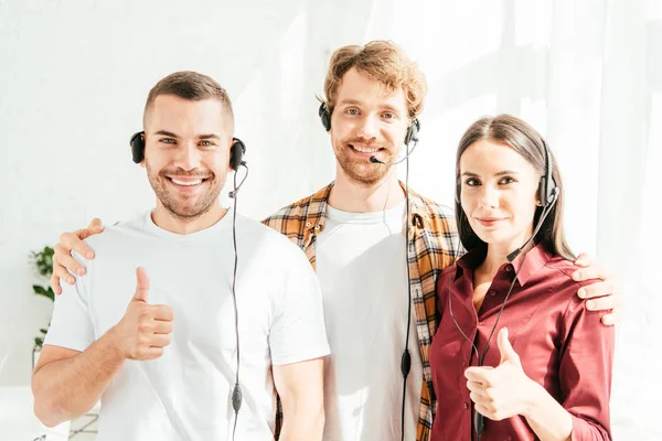 Happy brokers in headsets looking at camera and showing thumbs up — Stock Photo