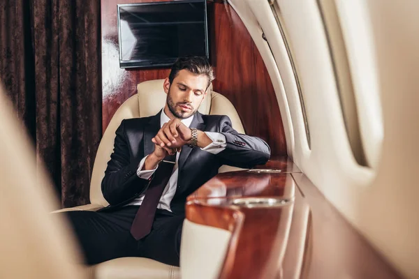 Красивый бизнесмен в костюме смотрит на наручные часы в частном самолете — Stock Photo