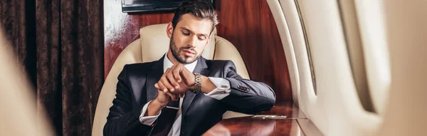 Panoramic shot of handsome businessman in suit looking at wristwatch in private plane — Stock Photo