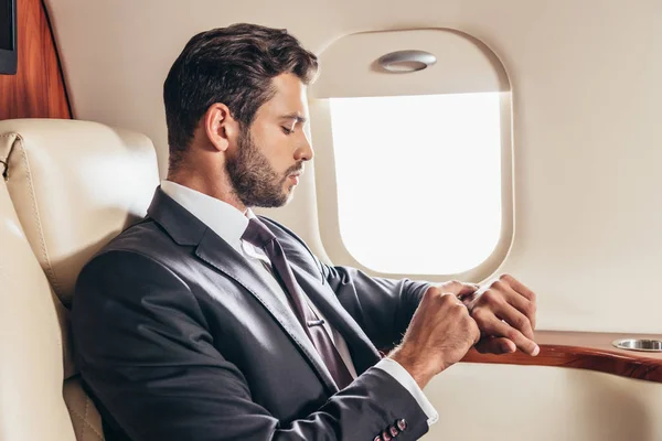 Vista lateral de hombre de negocios guapo en traje mirando reloj de pulsera en avión privado - foto de stock