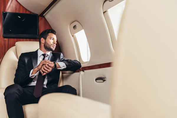 Selective focus of handsome businessman in suit looking through window in private plane — Stock Photo