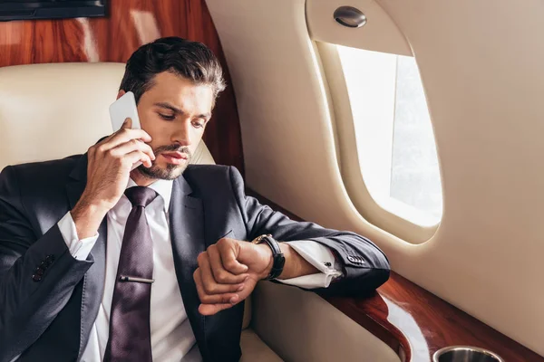 Handsome businessman in suit talking on smartphone and looking at wristwatch in private plane — Stock Photo