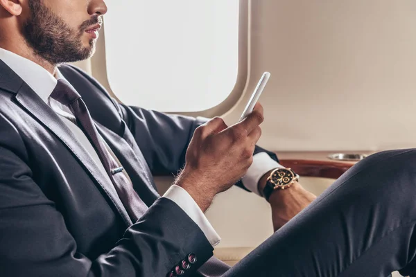 Cropped view of businessman in suit using smartphone in private plane — Stock Photo